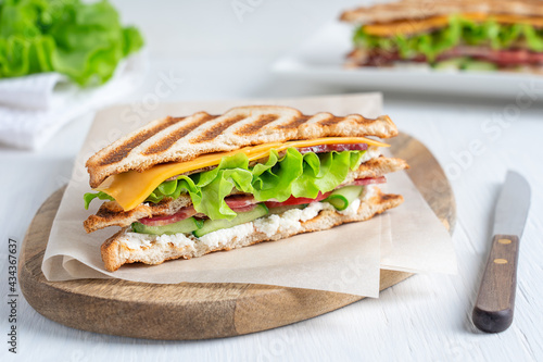 Homemade organic juicy sandwich made of ham, cheese, lettuce and tomatoes between slices of grilled toasted bread served on cutting board with knife on white wooden table at kitchen. Horizontal