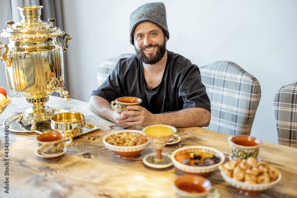 A portrait of the bathhouse attendant in bath hat sitting on the served table in russian style with samovar. Cheerful male personal in spa. A portrait of the bathhouse attendant in bath hat sitting on the served table in russian style with samovar. Cheerful male personal in spa.