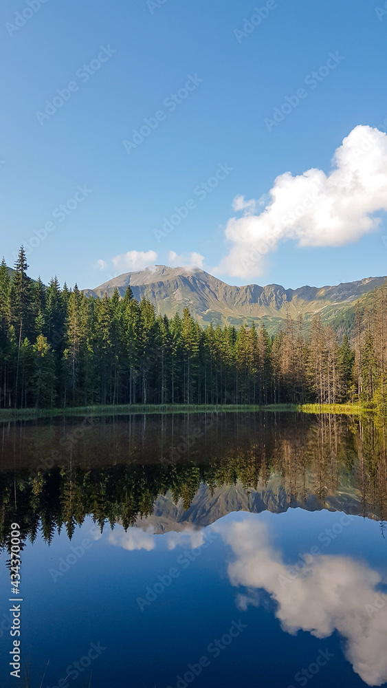 Fototapeta premium A panoramic view on Smreczynski Staw in Tatra Mountains in Poland. Glacial tarn at the mouth of Pysznianska Valley. The high Tatra chains are reflecting in the calm surface of the lake. White clouds