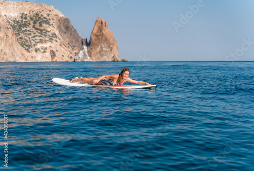 Sporty girl on a glanders surfboard in the sea on a sunny summer day. In a striped swimsuit, lying on his stomach, rowing with his hands. Summer activities by Stortom by the sea.