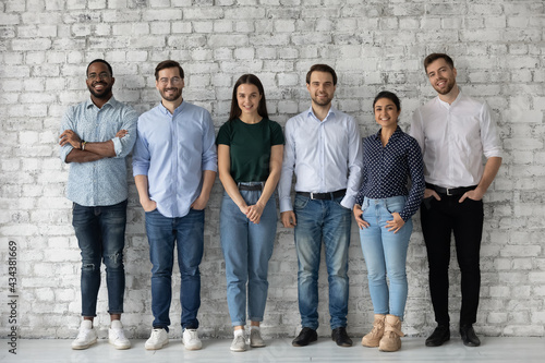 Group portrait of diverse millennial team of employees, company workforce, department staff. Happy multiethnic business people standing in row against wall, looking at camera, smiling. Full length