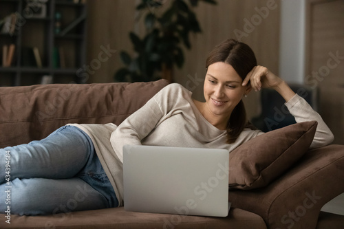 Interested millennial lady rest on couch with laptop read blog write comment to social network chat with mate online by wireless internet. Happy young female lying down on sofa working from home on pc