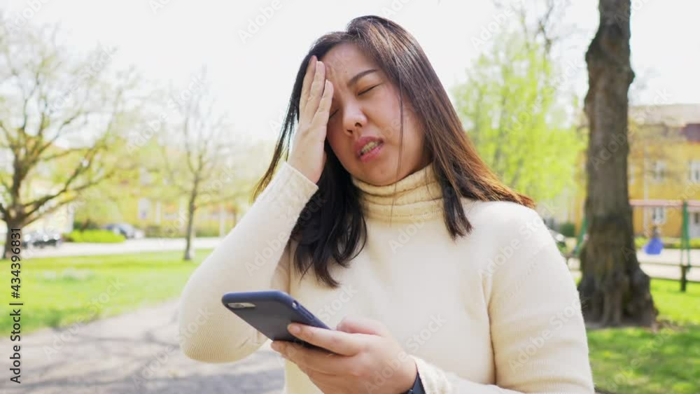 Asian black straight hair woman standing at park and using her phone to find something out. Boring face of Asian woman at park using social media Asian black straight hair woman standing at park and using her phone to find something out. Boring face of Asian woman at park using social media