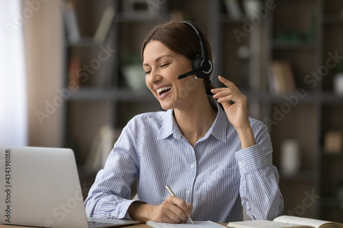 Funny remote learning. Joyful female student teenager taking notes of online virtual lesson laugh on good teacher speaker joke. Young woman in headset microphone having fun handwriting distant lecture