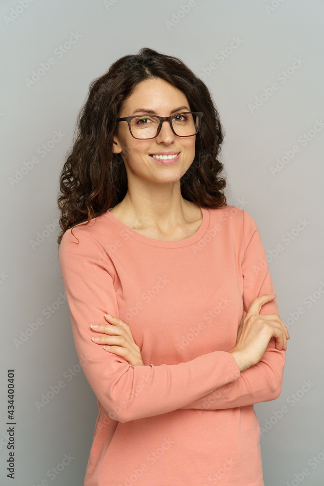 Fototapeta premium Portrait of smiling casual businesswoman standing with folded hands over gray wall. Confident business lady or teacher in eyeglasses and pink pullover. Millennial lady positive with curly black hair