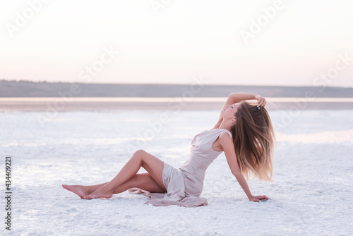 Young blonde woman in an evening airy pastel pink powdery dress sitting barefoot on white crystallized salt. Girl with natural make-up, hair is developing. Salt mining trip, walking on water at sunset