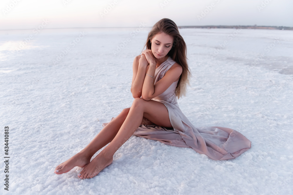 Young blonde woman in an evening airy pastel pink powdery dress sitting  barefoot on white crystallized