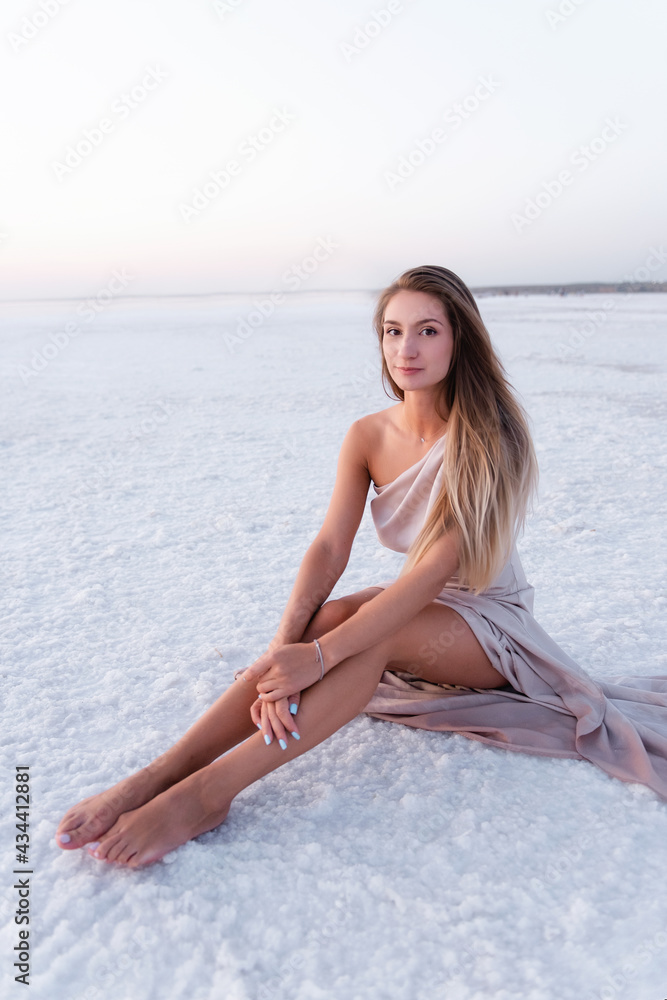 Young blonde woman in an evening airy pastel pink powdery dress sitting barefoot on white crystallized salt. Girl with natural make-up, hair is developing. Salt mining trip, walking on water at sunset