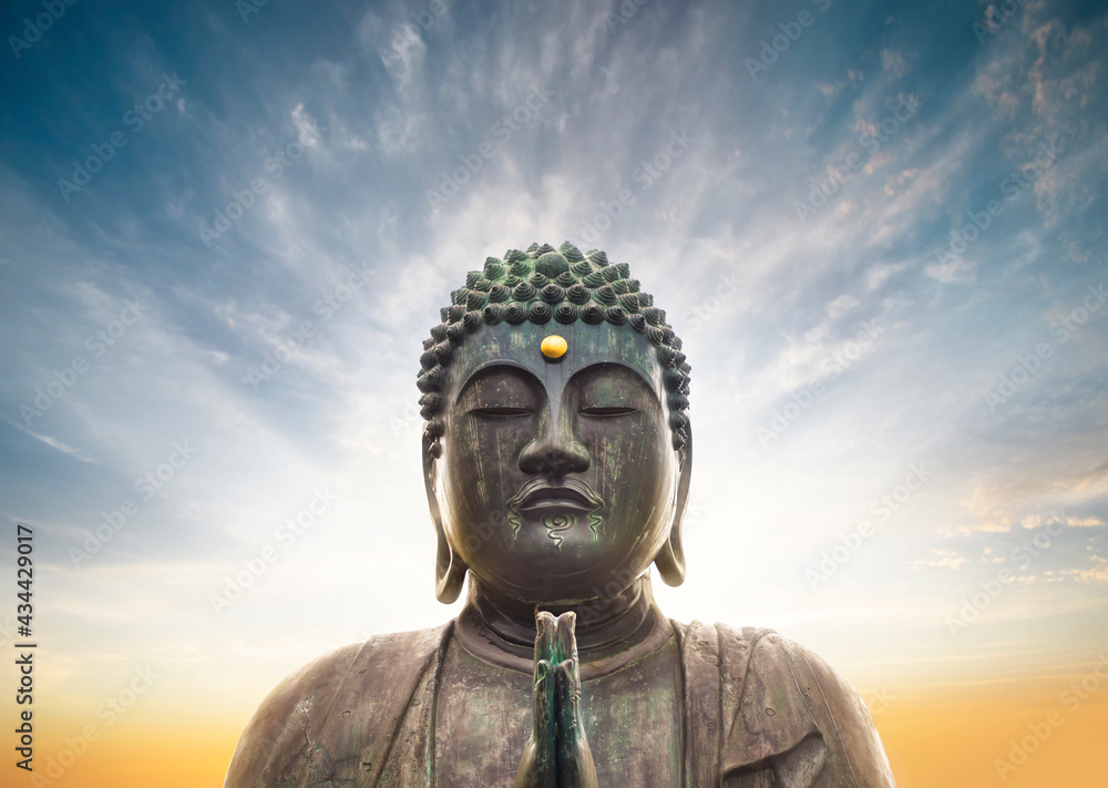 Bronze statue of Japanese Buddha Shaka Nyorai in meditation praying with two hands doing the gesture mudra called anjali surrounded by an aura glow on a sunset sky.