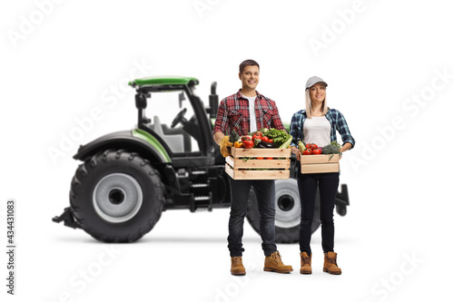 Young male and female farmers carrying crates with vegetables and posing in front of a tractor