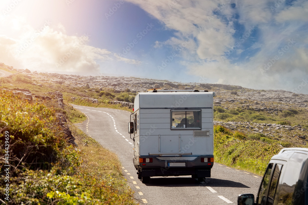 White camper van on a small asphalt road leading into sun. Traveling in motor home concept. Warm sunny day. Burren area, Ireland. Sun flare. Summer vacation concept