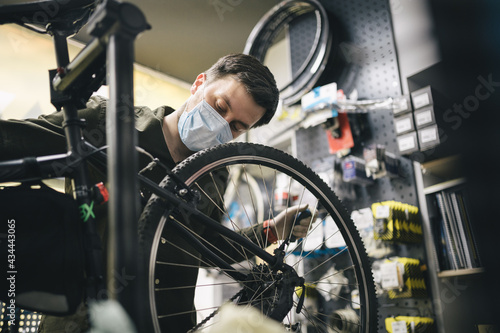 Bicycle mechanic wearing protective mask and gloves repairs customers bicycle wheel in accordance with quarantine standards during coronavirus pandemic. Small business during covid 19 lockdown