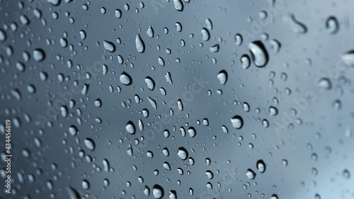 Water raindrop on a clear window glass. Raining on a car windscreen on a moody day. Moving droplets close up in front of a dark cloudy sky. Liquid fluid splashing on the windshield.