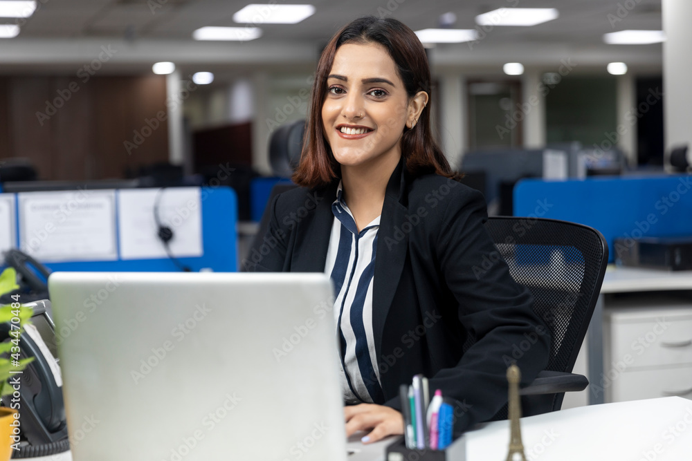Portrait of smiling pretty Indian businesswoman sitting at her workstation against office background, working on laptop, corporate environment,