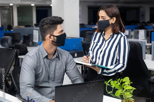 Portrait of two young Indian businesspeople wearing Covid protection mask and discussing project at workstation, corporate environment.
