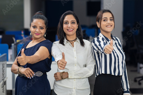 Three Indian businesswomen standing together against office background, showing thumbs up sign of victory, corporate environment.
