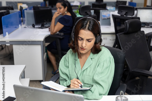 Pretty Indian businesswoman wearing headphones, sitting at her workstation, office background, looking into her diary, corporate environment.