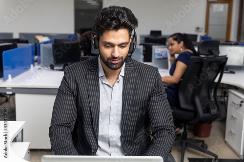 Portrait of young Indian businessman wearing headphones, sitting at his workstation against office background, corporate environment, call centre.