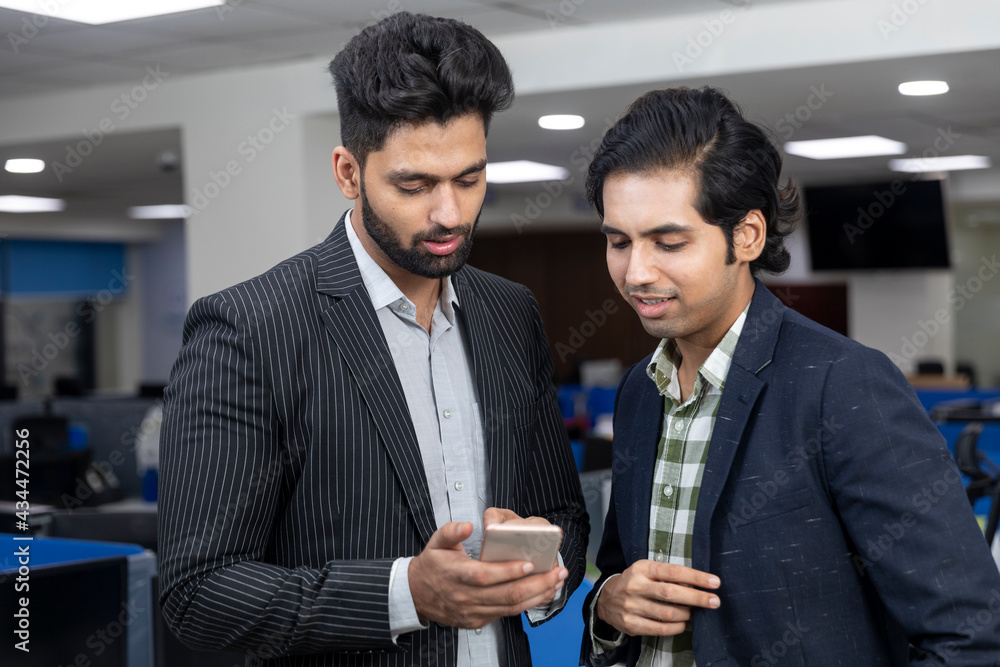 Portrait of two young handsome Indian businessmen looking into phone, discussing project, corporate environment.