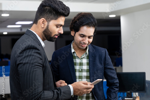 Portrait of two young handsome Indian businessmen looking into phone, discussing project, corporate environment.