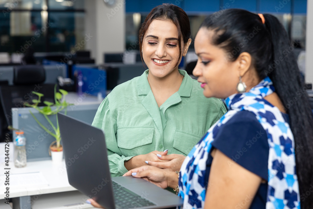 Portrait of two young Indian businesswomen looking into laptop, discussing project, corporate environment, office background. Portrait of two young Indian businesswomen looking into laptop, discussing project, corporate environment, office background.
