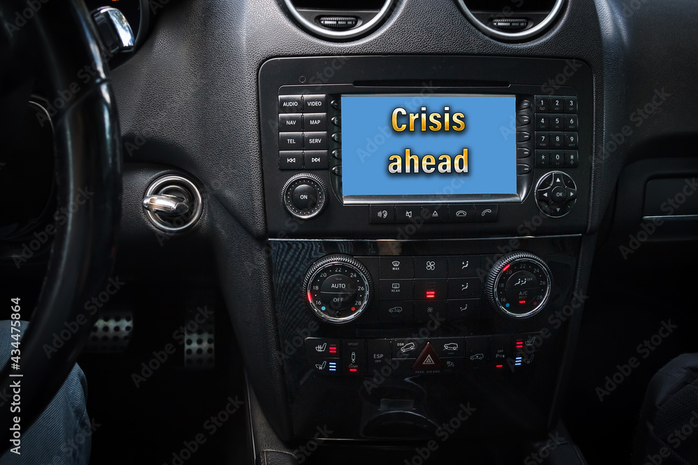 CRISIS AHEAD is written on the dashboard of the car next to the controls, ignition key and steering wheel