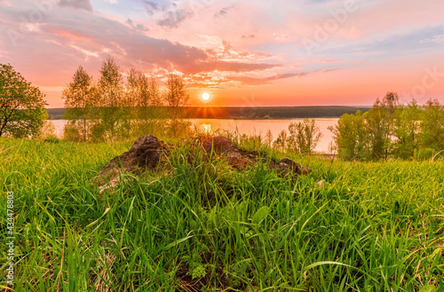 Scenic view at beautiful sunset on a shiny lake with old rough stone on the foreground  green grass  birch trees  golden sun rays  calm water  nice cloudy sky on a background  spring landscape