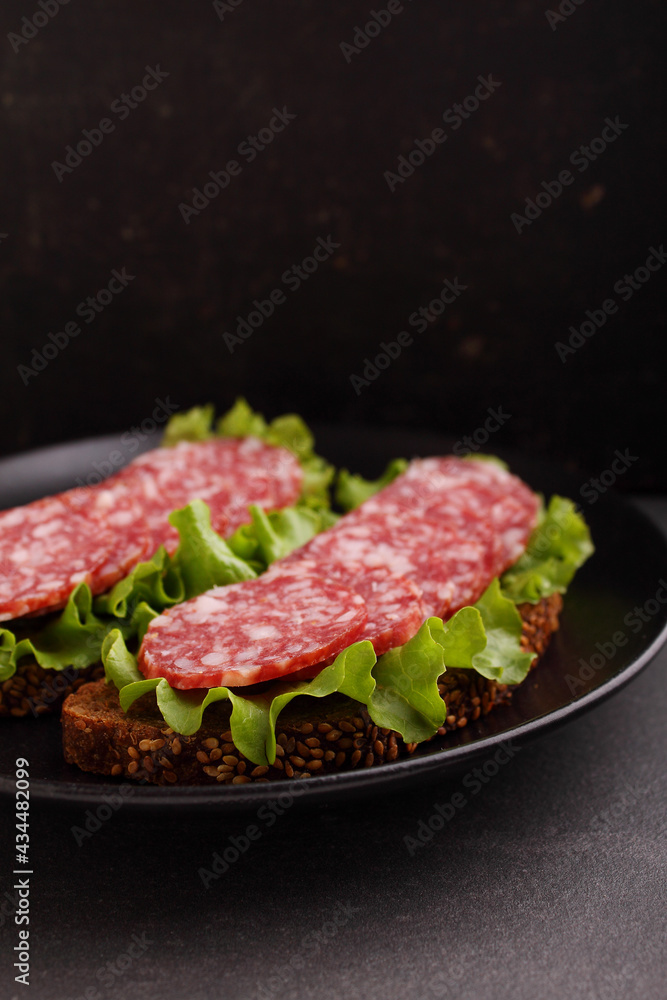 Delicious sandwich on black grain bread with lettuce and smoked sausage on plate on black background