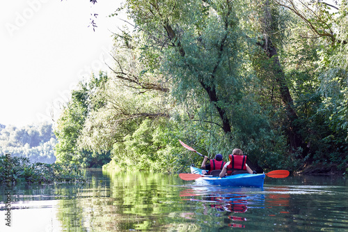 Mother and son paddle blue kayak near green trees on wildlife areas of Danube river. Summer kayaking. Back view. Concept for family, adventure, travel, action, lifestyle and kayaking