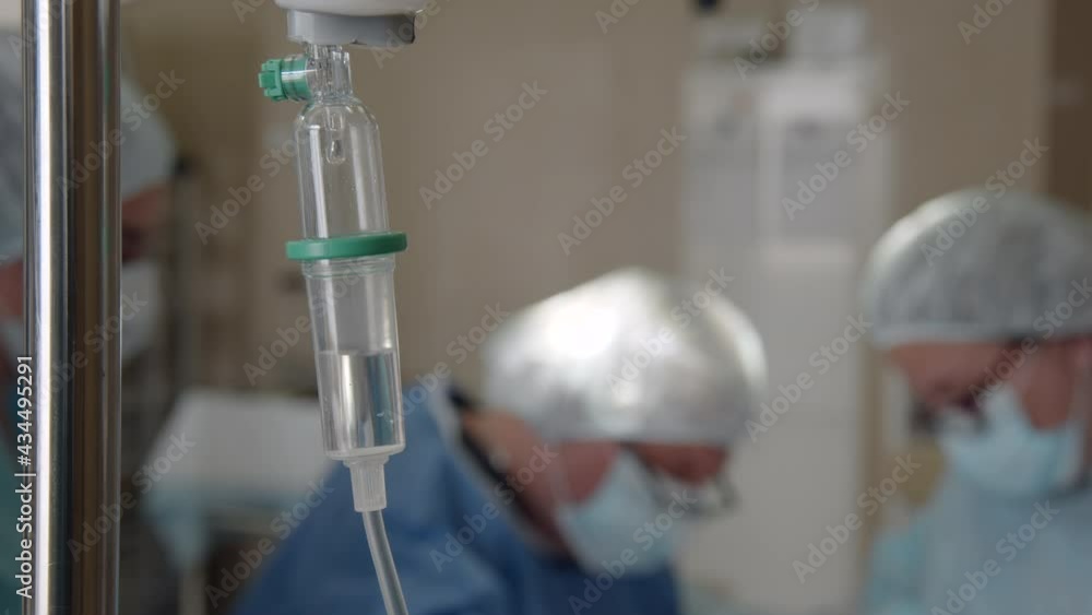 The solution for anesthesia of the patient during the operation is dripped into the dropper. In the background, surgeons in poppies, gowns and medical hats and glasses perform an operation on a person The solution for anesthesia of the patient during the operation is dripped into the dropper. In the background, surgeons in poppies, gowns and medical hats and glasses perform an operation on a person