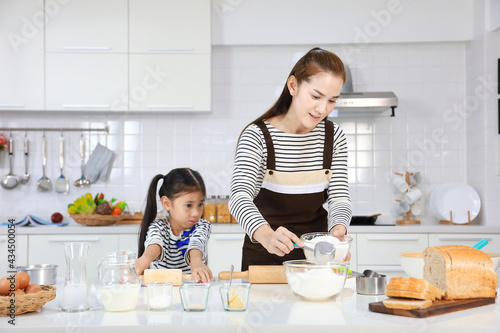 Happy Asian mother teaching her young daughter to bread baking in white modern kitchen while mixing recipe for kneading flour to make dough