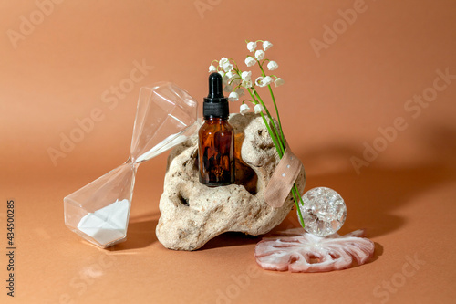 Modern still life with serum bottle on background of unusual natural stone, with bouquet of lilies of the valley and hourglass. The concept of anti-aging care products photo