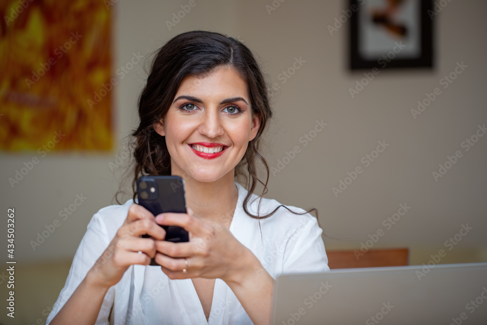 Businesswoman smiling to the camera from home office. Happy female emplyee working from home with positive mindset. Satisfied young adult feminine holding mobile in hands. Businesswoman smiling to the camera from home office. Happy female emplyee working from home with positive mindset. Satisfied young adult feminine holding mobile in hands.