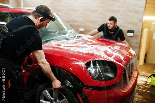 Car service worker put anti gravel film on a red car body at the detailing vehicle workshop. Car protection with special films. © AS Photo Family