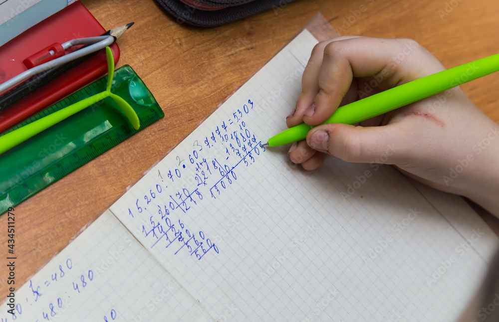 A girl's hand with a ballpoint pen solve an example in mathematics close-up. The student performs the task in the notebook. The student does his homework at the table. Warm soft daylight. A girl's hand with a ballpoint pen solve an example in mathematics close-up. The student performs the task in the notebook. The student does his homework at the table. Warm soft daylight.