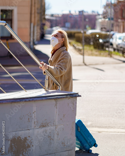 Fashionable senior woman traveler wearing respirator or face mask standing on sunny street with blue suitcase and looking up. Elderly lady getting lost in city while traveling during covid19 outbreak