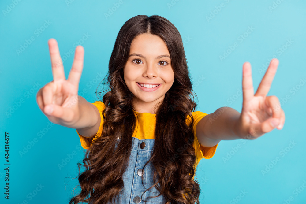 Photo of adorable cute small school girl wear denim jeans overall smiling showing two v-signs isolated blue color background Photo of adorable cute small school girl wear denim jeans overall smiling showing two v-signs isolated blue color background