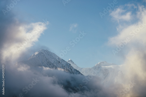 Beautiful view of snow-capped mountains above thick clouds in sunshine. Scenic bright mountain landscape with white-snow peak among dense low clouds in blue sky. Wonderful scenery with snowy pinnacle. © Daniil