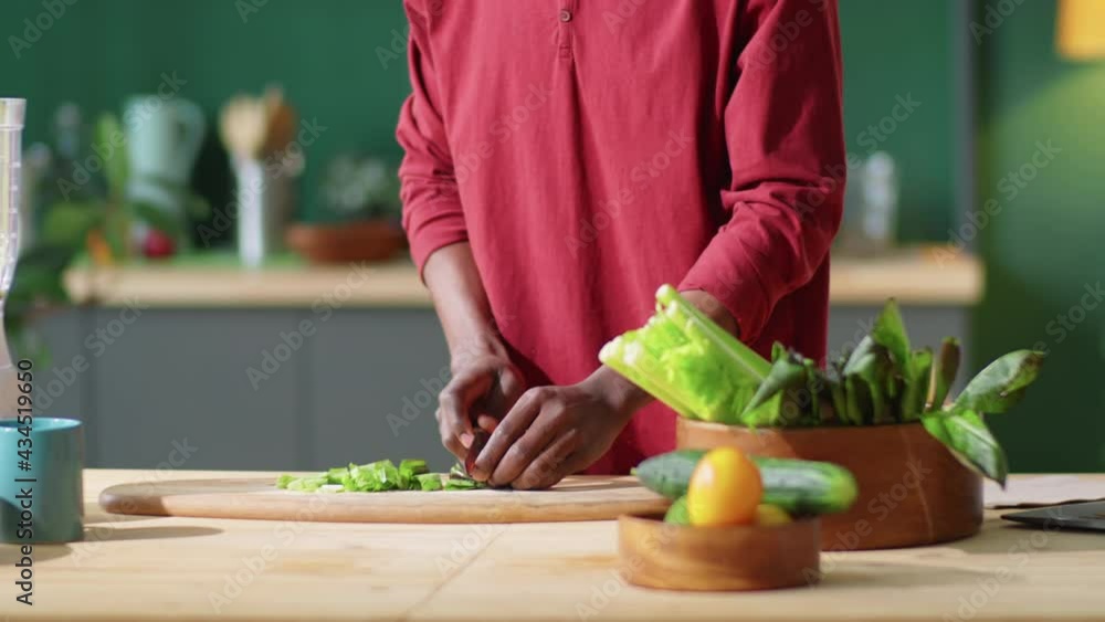 Tilt down shot of young Afro-American man cutting green salad leaves and putting them into blender while preparing healthy food in kitchen at home Tilt down shot of young Afro-American man cutting green salad leaves and putting them into blender while preparing healthy food in kitchen at home