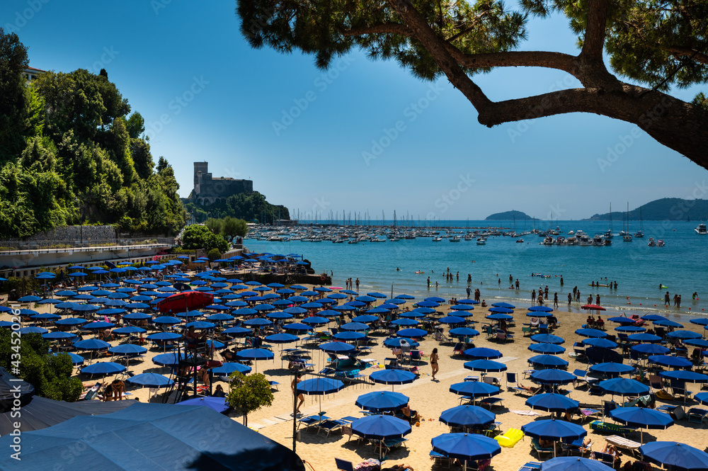 Lerici, Liguria, Italy. June 2020. The silhouette of a pine trunk with the background of the castle of Lerici frame the beach full of blue umbrellas and people. Beautiful sunny summer day.