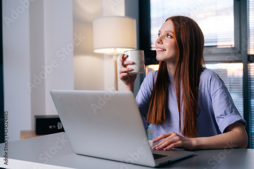 Dreamy thinking young woman using laptop computer and drinking coffee at home workplace. Business lady watching news at laptop screen in creative office. Female professional working on notebook.