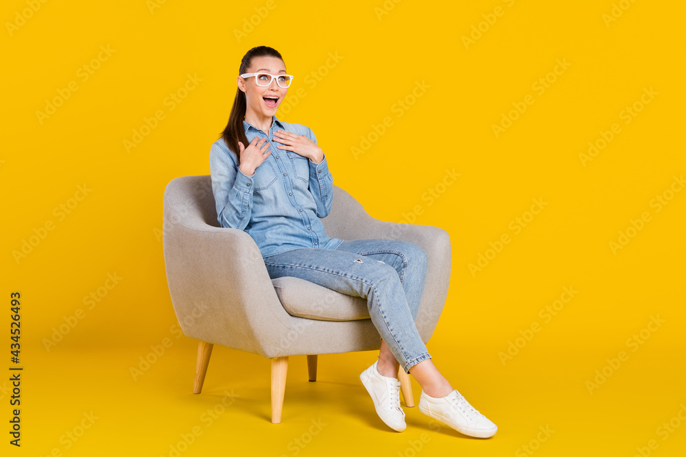 Full length body size photo woman sitting in chair amazed keeping hands on chest in glasses isolated vibrant yellow color background Full length body size photo woman sitting in chair amazed keeping hands on chest in glasses isolated vibrant yellow color background