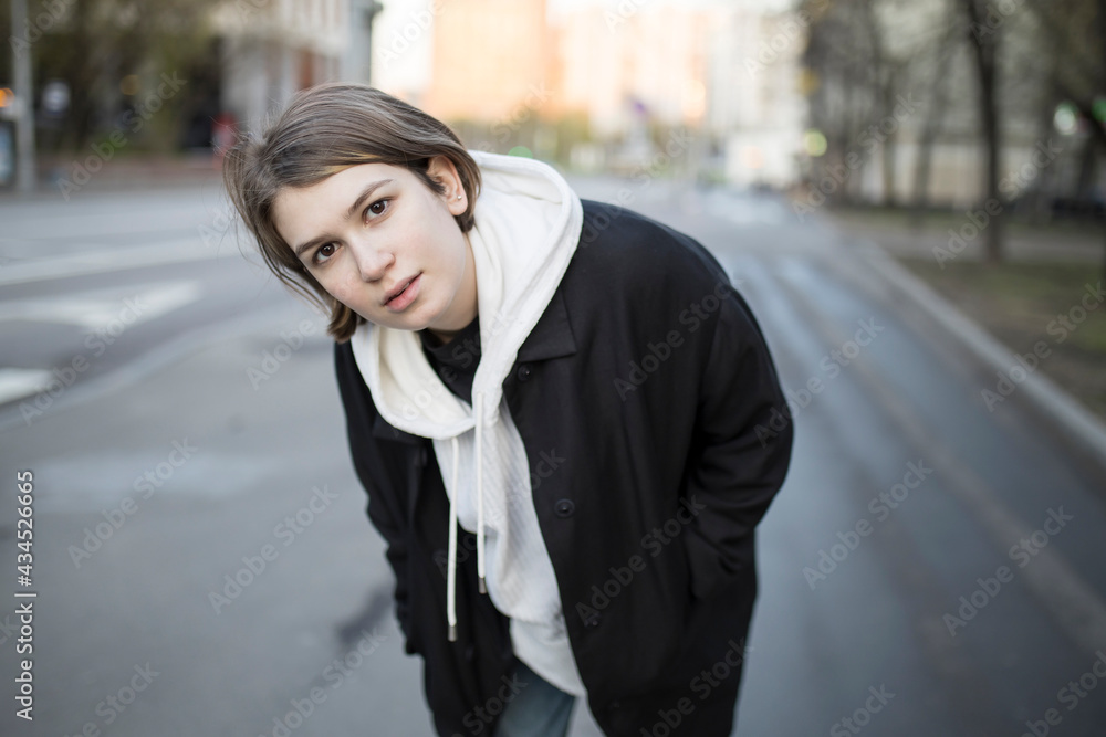 A serious short-haired girl in a white hoodie with a hood, a black raincoat and jeans walks down the street. A serious short-haired girl in a white hoodie with a hood, a black raincoat and jeans walks down the street.