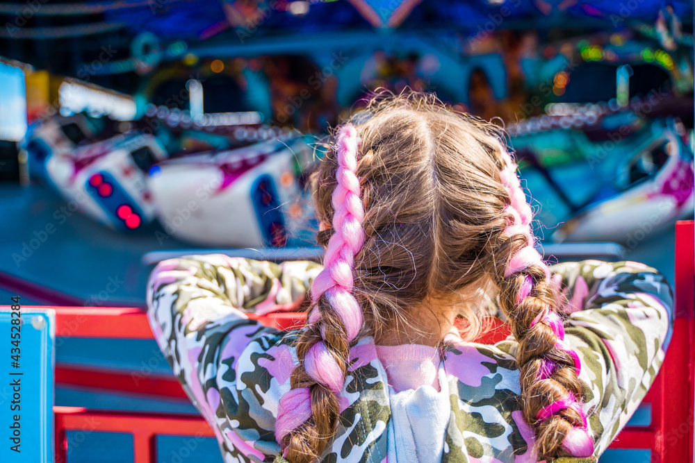 Rear view of a little girl with colorful hair looking at an amusement park ride. Kazan, Russia - 8 May 2021 Rear view of a little girl with colorful hair looking at an amusement park ride. Kazan, Russia - 8 May 2021