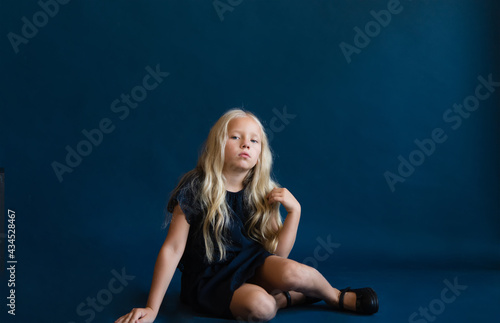 beautiful blonde in a dark blue dress on a solid blue background. The photo is great for demonstrating the character of a child - daydreaming, enthusiasm, vulnerability, touching, beauty, stubbornness