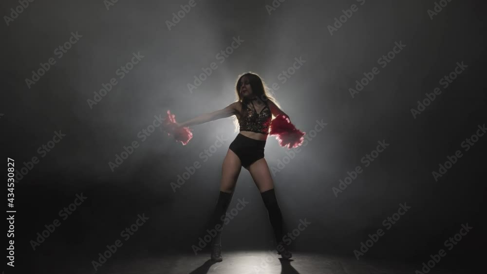 Young female cheerleader with orange pompoms in uniform is dancing on black smoky studio background with backlight. Silhouette of beautiful dancer performs cheering dance. Slow motion ready 59.94fps. Young female cheerleader with orange pompoms in uniform is dancing on black smoky studio background with backlight. Silhouette of beautiful dancer performs cheering dance. Slow motion ready 59.94fps.