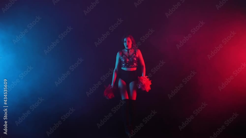 Young cheerleader with pompoms in uniform is dancing on black smoky studio background with red and blue lights. Silhouette of beautiful dancer performs cheering dance. Slow motion ready 59.94fps. Young cheerleader with pompoms in uniform is dancing on black smoky studio background with red and blue lights. Silhouette of beautiful dancer performs cheering dance. Slow motion ready 59.94fps.
