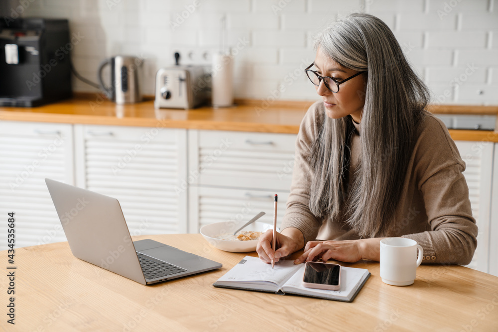 Mature grey woman writing down notes while working with laptop Mature grey woman writing down notes while working with laptop