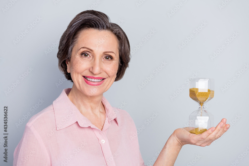 Photo of confident pretty age woman dressed pink shirt smiling holding hourglass isolated grey color background Photo of confident pretty age woman dressed pink shirt smiling holding hourglass isolated grey color background