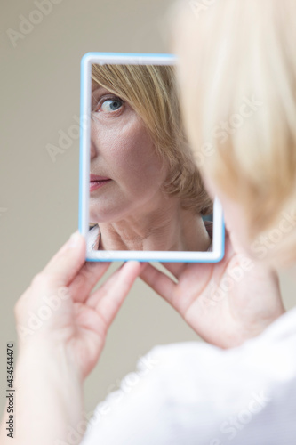 Portrait of Caucasian Matre Woman Examining Her Face In Hand Mirror Indoors